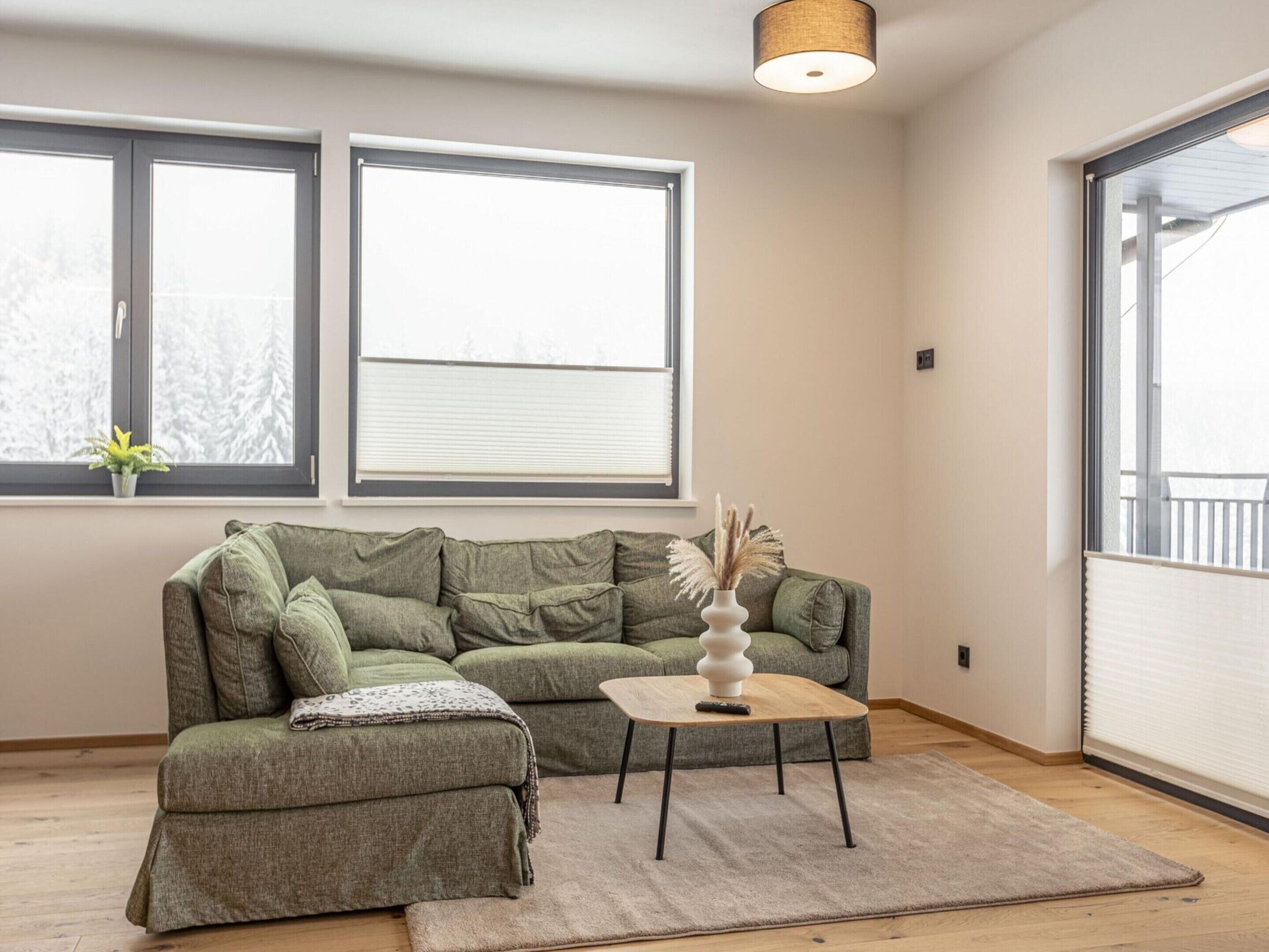 A living room with a sectional sofa, a small coffee table, and a rug. There are two windows with blinds, and a ceiling light fixture.