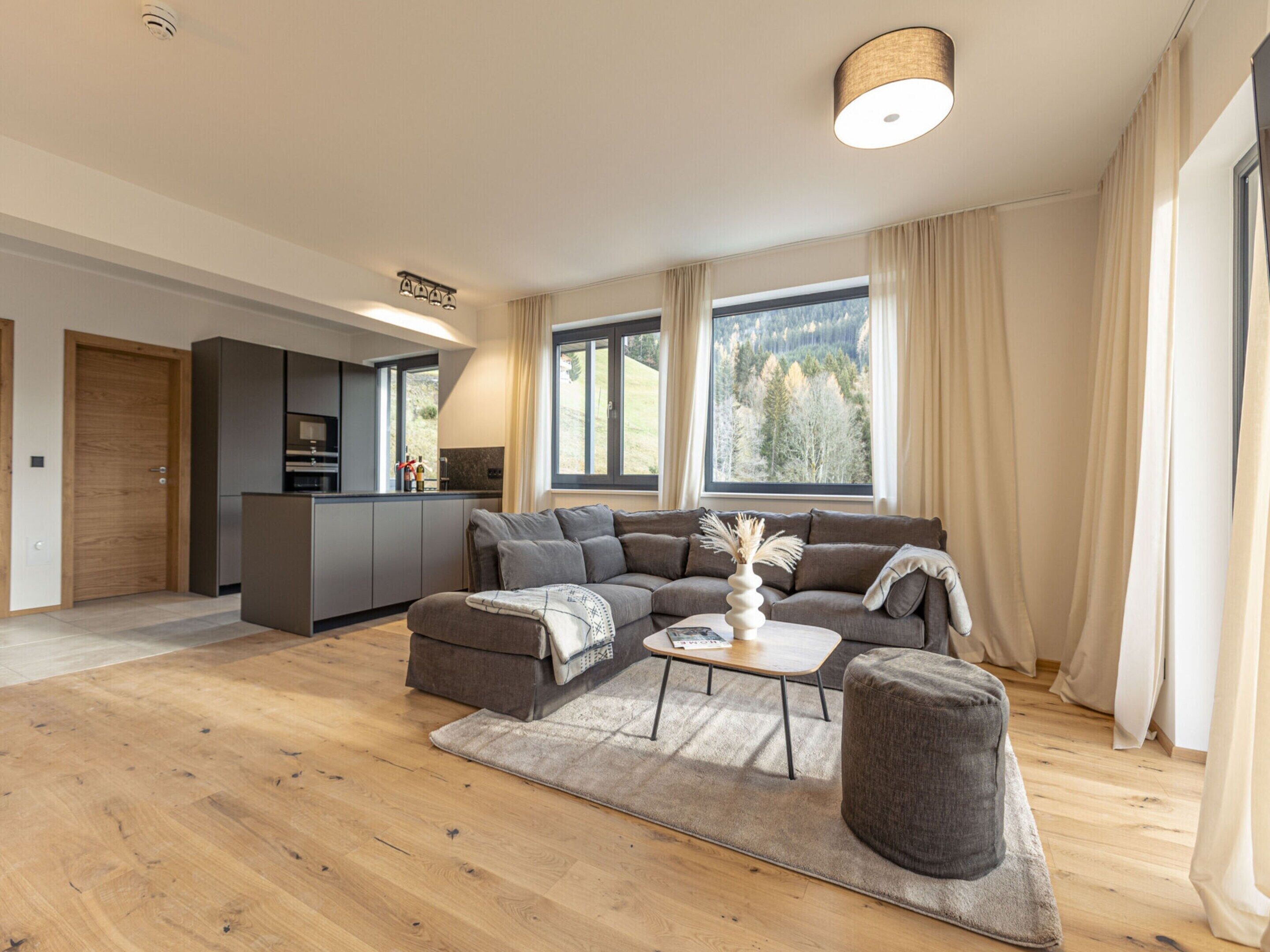 A modern living room with a sofa, ottoman, and coffee table, a kitchen area, and large windows with curtains.