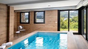 Una piscina interna moderna con pareti in legno e ampie finestre che offrono una vista panoramica su colline e un lago.