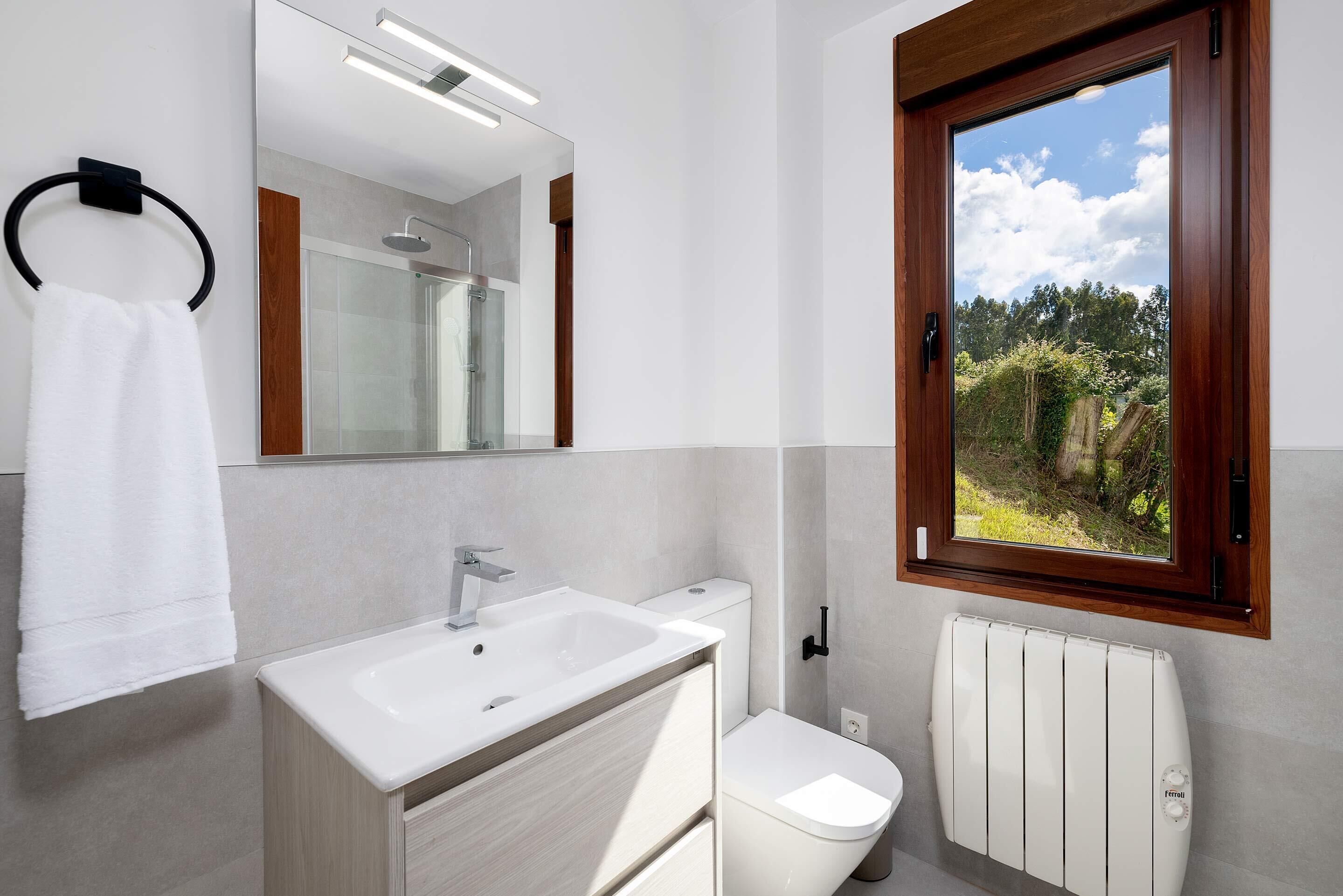 Un baño moderno con lavamanos, inodoro y radiador blancos, un espejo grande y un ventanal con vistas a zonas verdes.