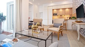Ein modernes Wohnzimmer mit Glastisch, zwei Sesseln, einer Couch, einer Küchenzeile mit Holzschränken und einem an der Wand montierten Fernseher.