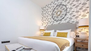Ein ordentlich eingerichtetes Schlafzimmer mit einem großen Bett, zwei Nachttischen mit Lampen, einem wandmontierten runden Spiegel und einem Wandpapier mit Muster.