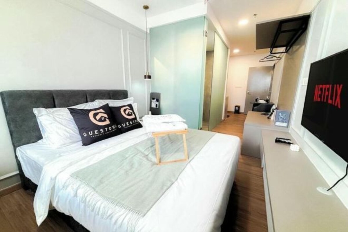 A hotel room with a bed, a small wooden table, a TV, and a wall-mounted shelf.