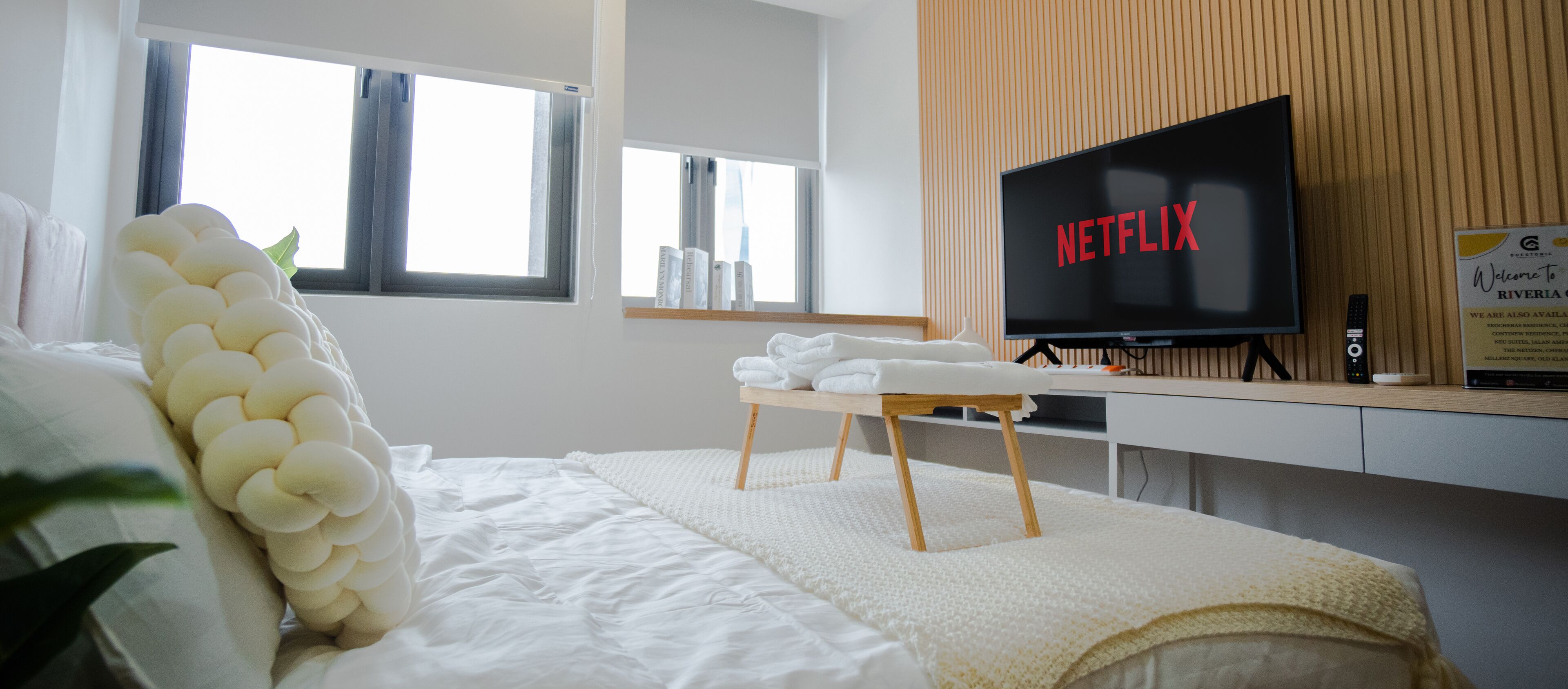 A hotel room with a bed, a bedside table with towels, a television displaying 'NETFLIX', and a window with curtains.