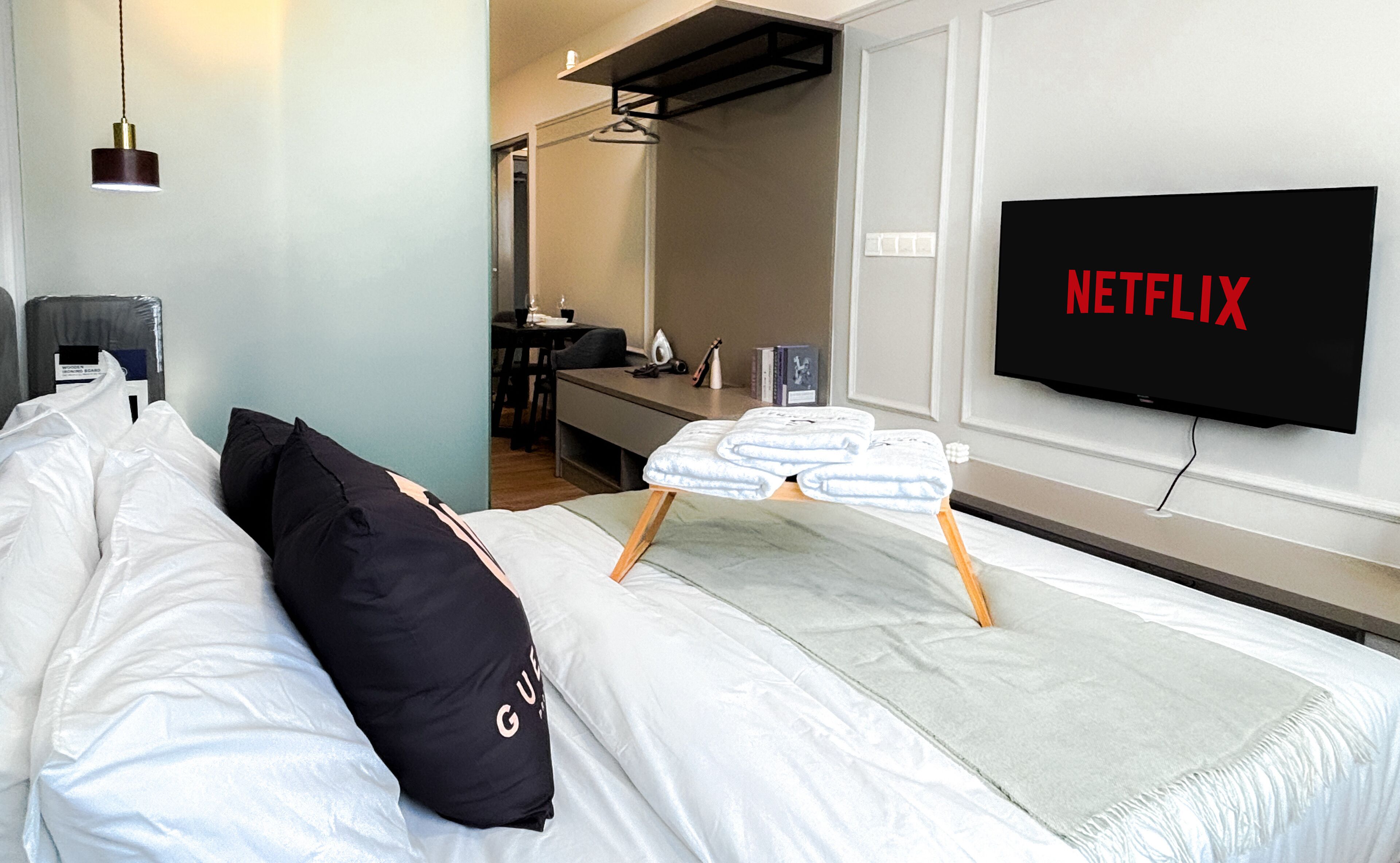 A hotel room with a bed, a TV mounted on the wall displaying 'NETFLIX', a small table with a chair, and a desk with a chair.