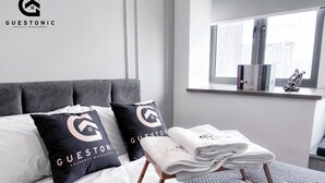 A modern hotel room with a grey upholstered headboard, white bedding, and a grey textured rug. There is a window with a view of buildings and a shelf with books and a guitar. - Riveria City KL Sentral by Guestonic (Kuala Lumpur)