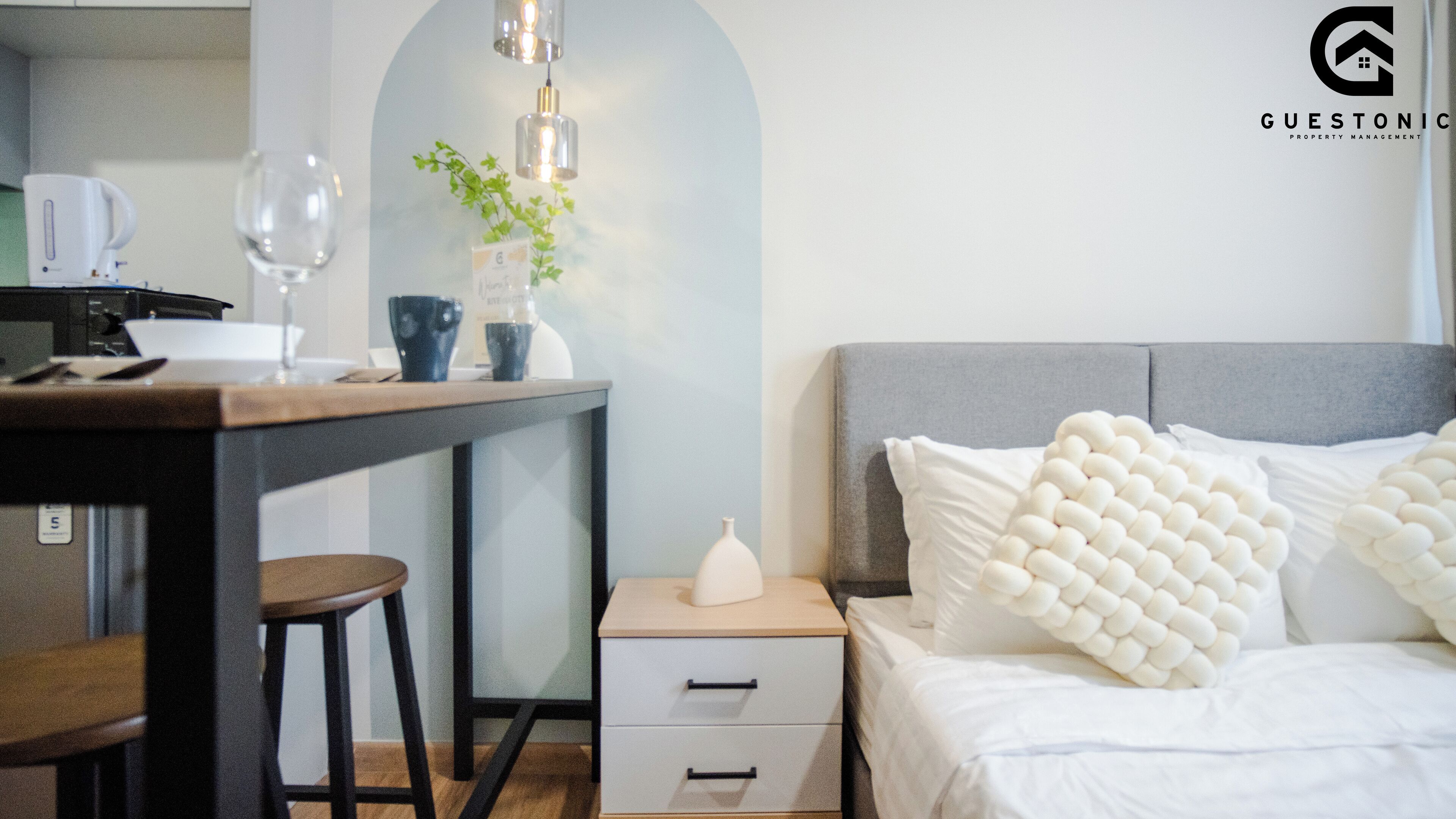 A modern hotel room with a wooden desk, a single bed with a grey headboard, and a small bedside table with a white vase.
