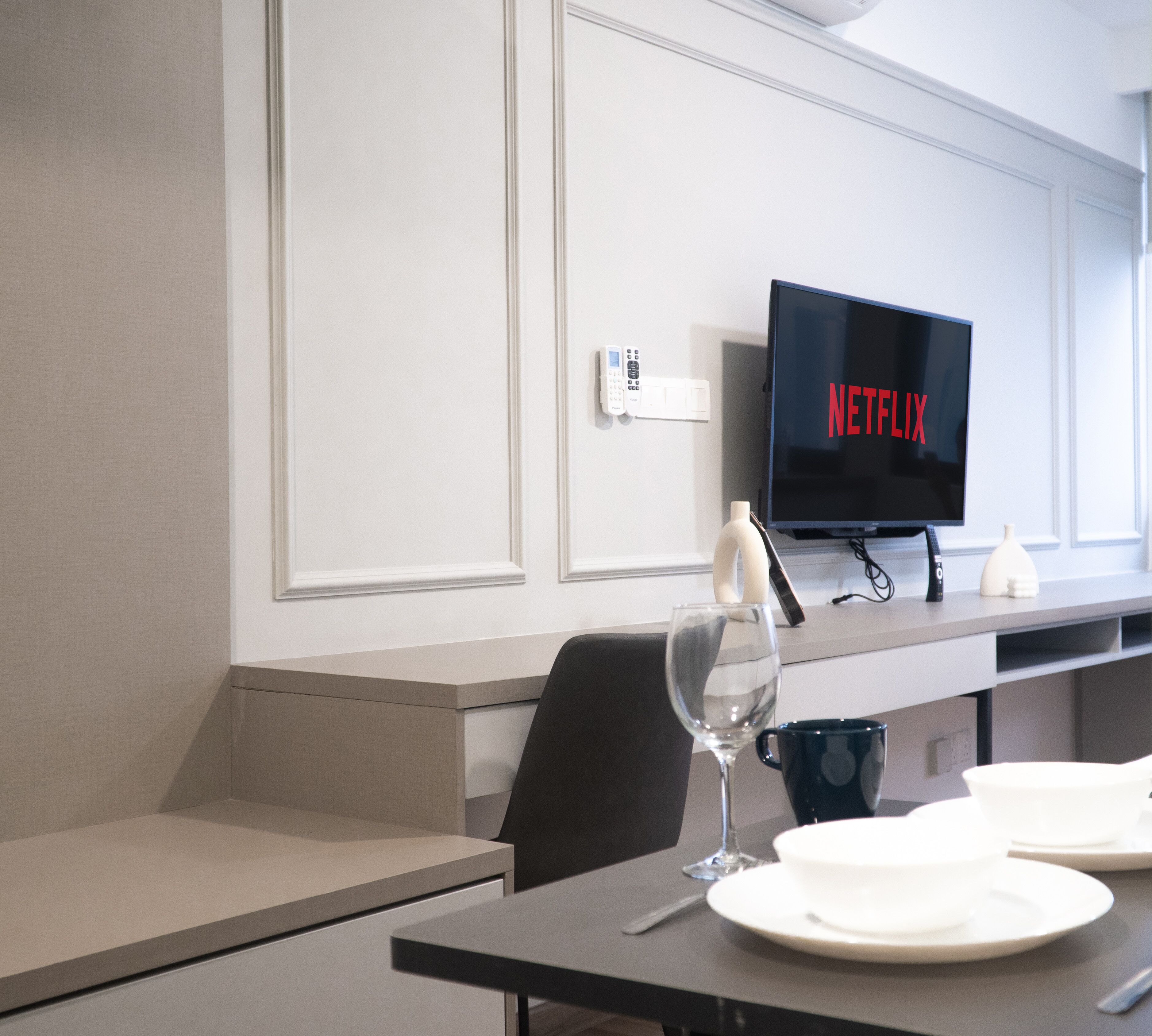 A modern hotel room with a dining table set for two, a wine glass, a remote control, and a television displaying the Netflix logo.
