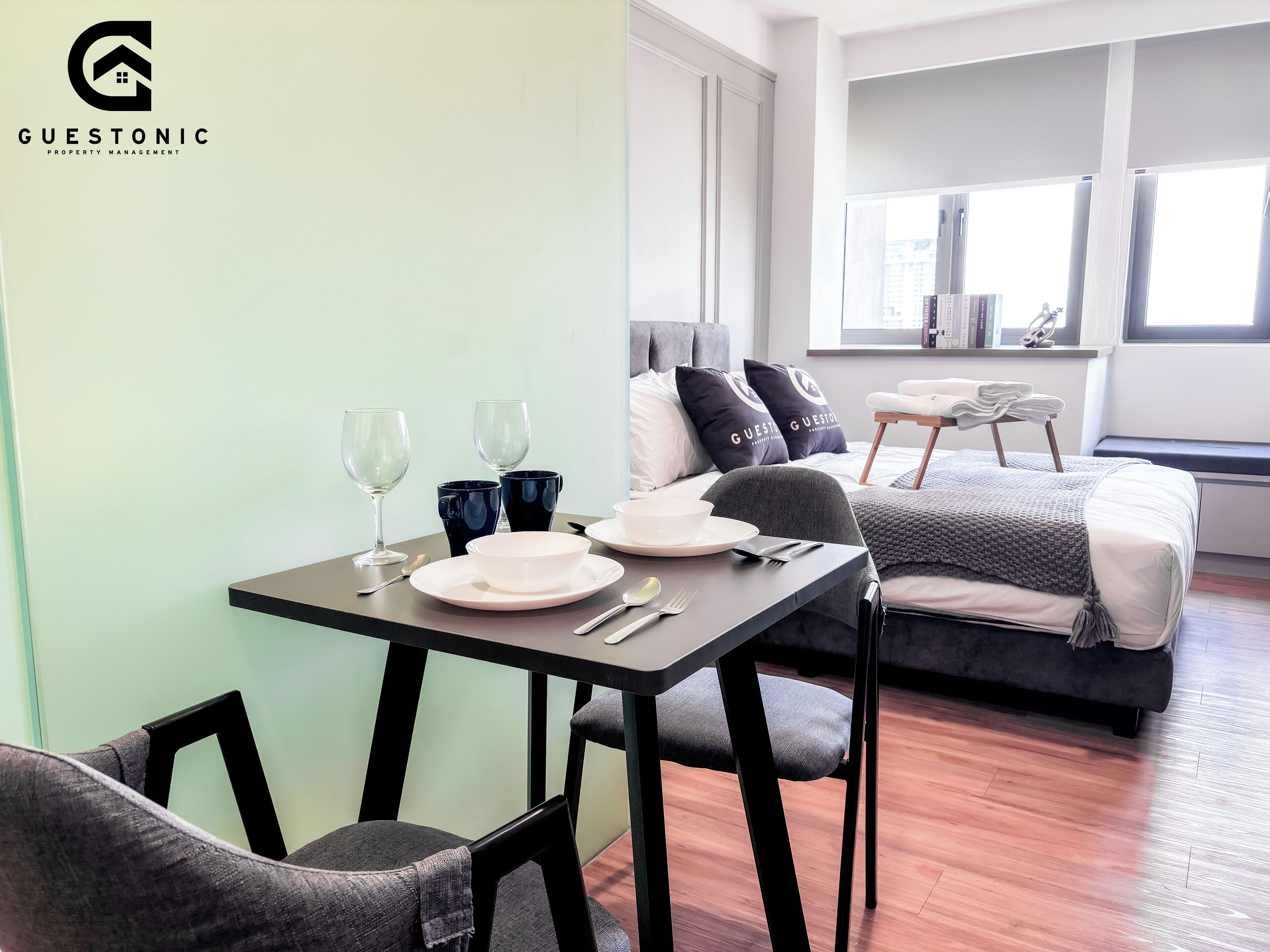 A modern, well-lit room with a dining table set for two, a grey sofa, and a bed with a grey blanket.