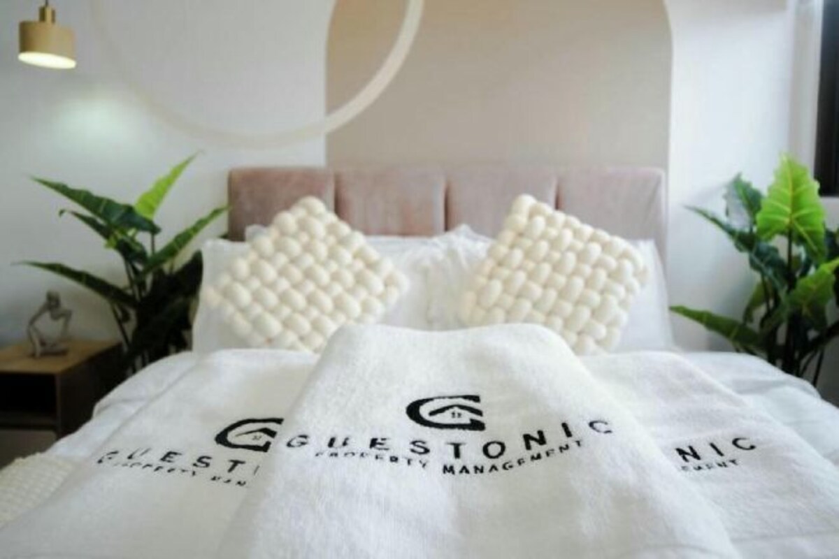 A bed with a white blanket and a logo, a headboard, and two bedside plants.