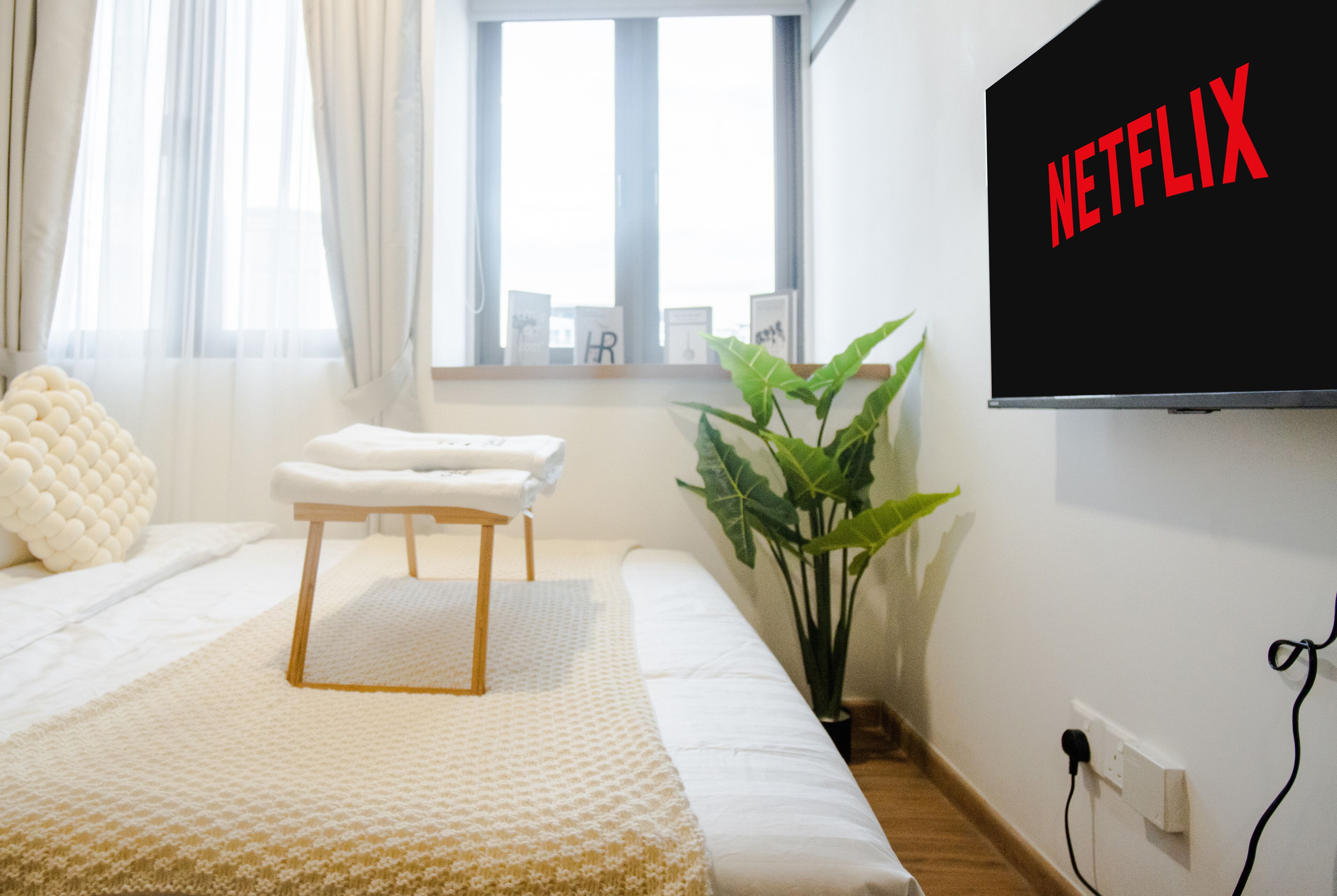 A neatly arranged bedroom with a bed, a bench, a potted plant, and a wall-mounted TV displaying the Netflix logo.