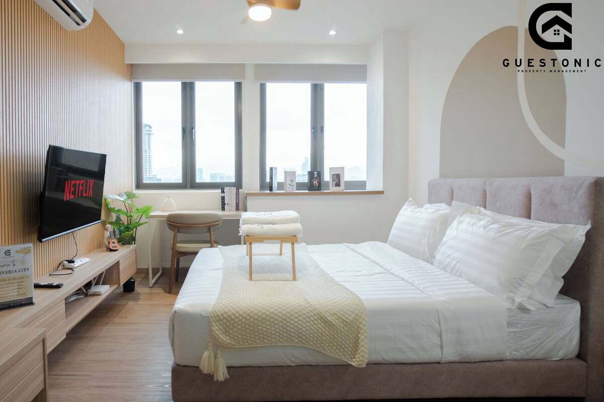 A modern hotel room with a bed, a desk with a computer, a TV, and a window with a city view.
