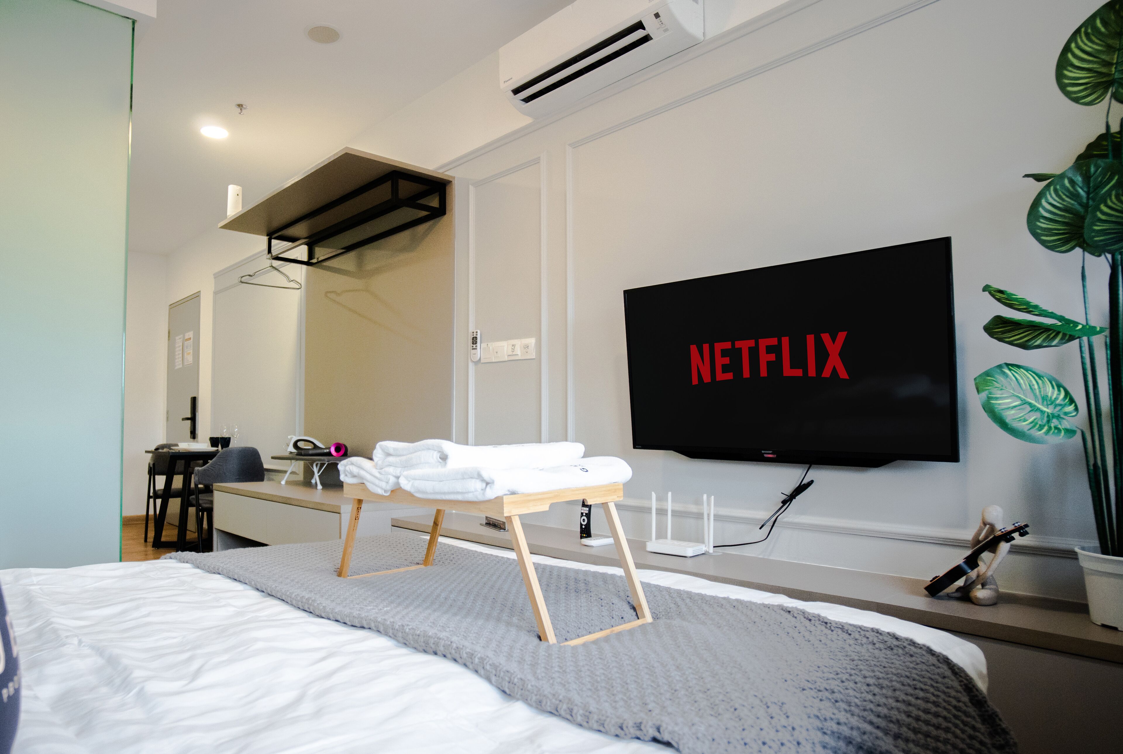 A modern hotel room with a bed, a desk, a chair, a TV mounted on the wall, and a plant.