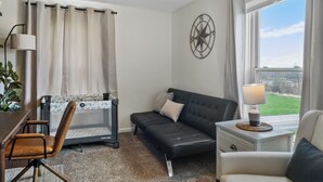 A room with a desk, a cushioned chair, a sofa, a small table with a lamp, and a window with curtains.