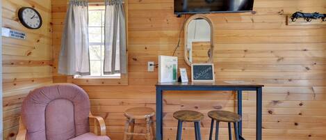 Ein Holzblockhaus-Interieur mit einem gepolsterten Sessel, einem kleinen Tisch mit Hockern, einer Wanduhr und einem angebrachten Fernseher.