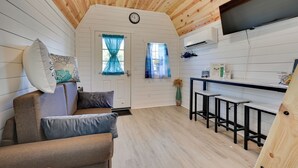 Een gezellig interieur van een chalet met een houten plafond, een bank met kussens, een keuken met een barkruk en een aan de muur bevestigde televisie.