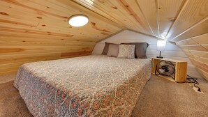 Een knus slaapkamer op zolder met een houten schuine dak, een bed met beddengoed met een patroon, een bedlampje en een draagbare ventilator.