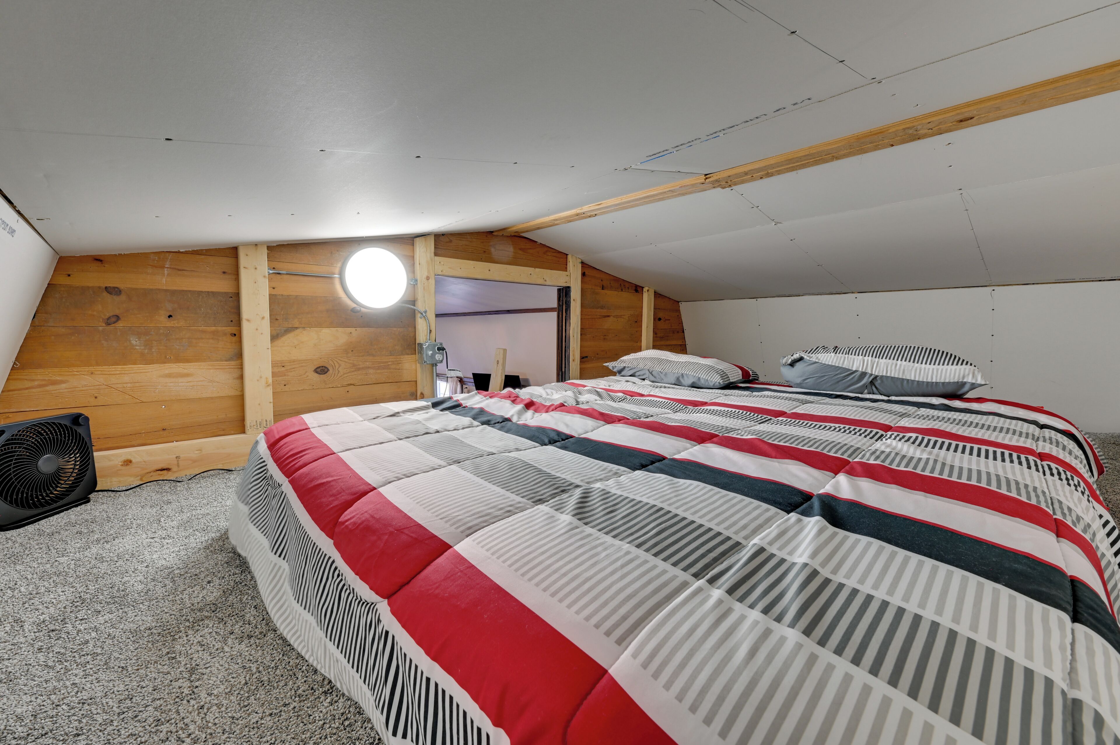 Une chambre avec un plafond en pente, un mur en bois, un lit avec une literie à rayures, un ventilateur et une petite fenêtre.
