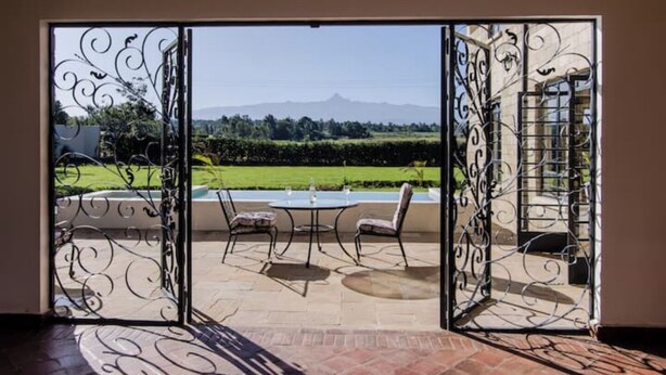 Una puerta corrediza de vidrio da a un patio exterior con mesa y sillas, piscina y vistas a un paisaje verde y montañas.