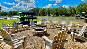 A lakeside deck with Adirondack chairs, a fire pit, and a green boat docked by the water. - LOWER Level of "Your Big Lake House" - Firepit, Kayaks, HotTub, etc.  (Cedar Bluff)