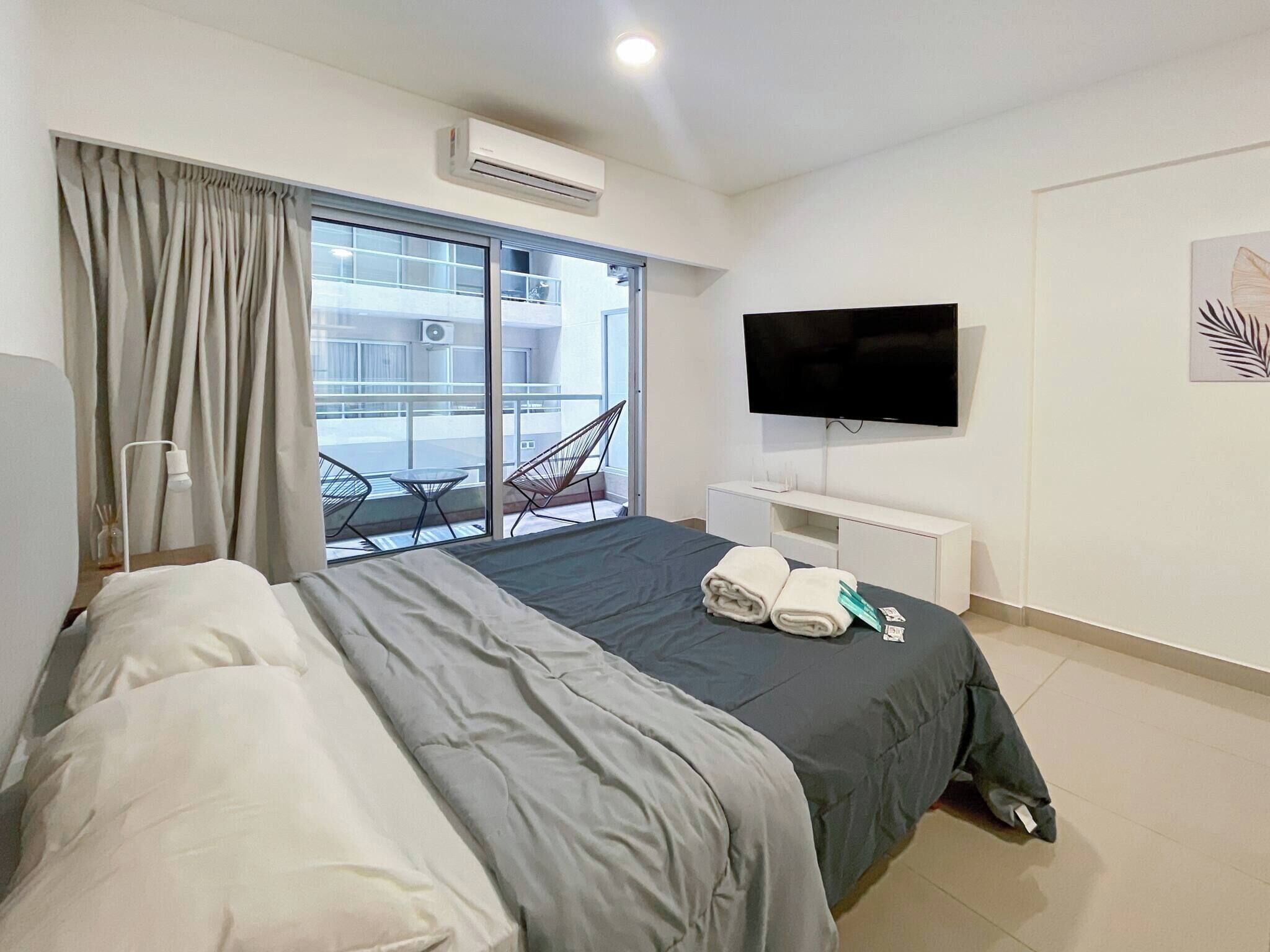 A hotel room with a bed, a TV, a window with curtains, a balcony with chairs, and an air conditioning unit.