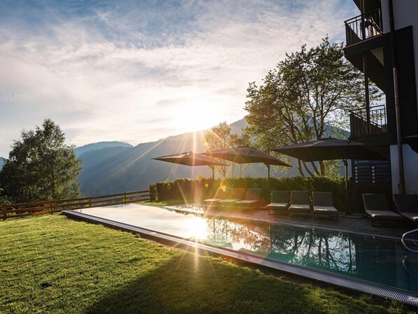 Ein Pool mit Liegestühlen und Sonnenschirmen, umgeben von einem Holzzaun und üppiger Vegetation, mit Bergen im Hintergrund.