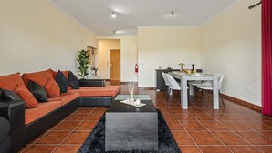 A modern living room with a sectional sofa, a dining table with chairs, and a kitchen area in the background. - Palm Tree House by Homie (São Vicente)