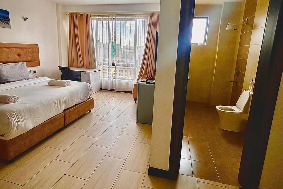A hotel room with a bed, a desk, a chair, a window with curtains, and a bathroom visible through an open door.