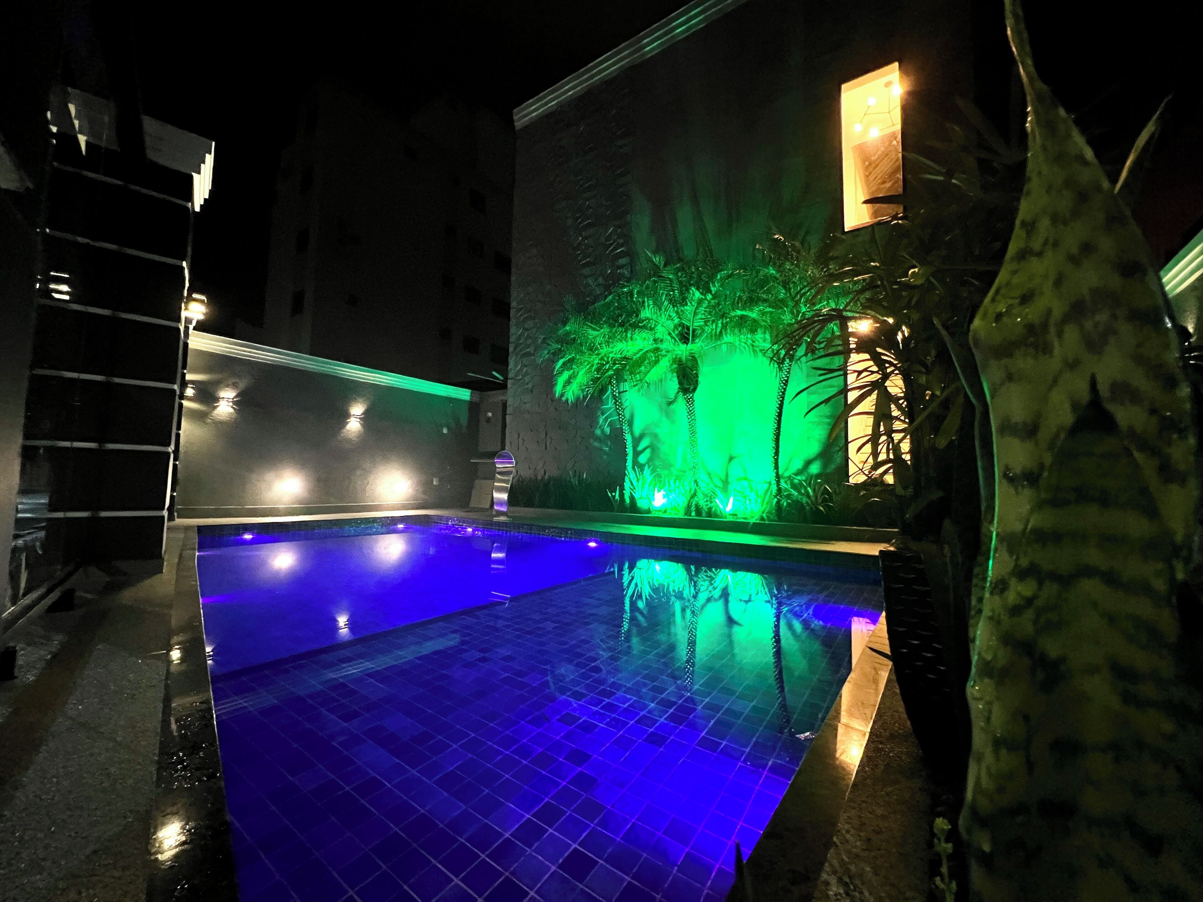 Una piscina con iluminación azul, rodeada por un muro con un diseño de palmera iluminada en verde.