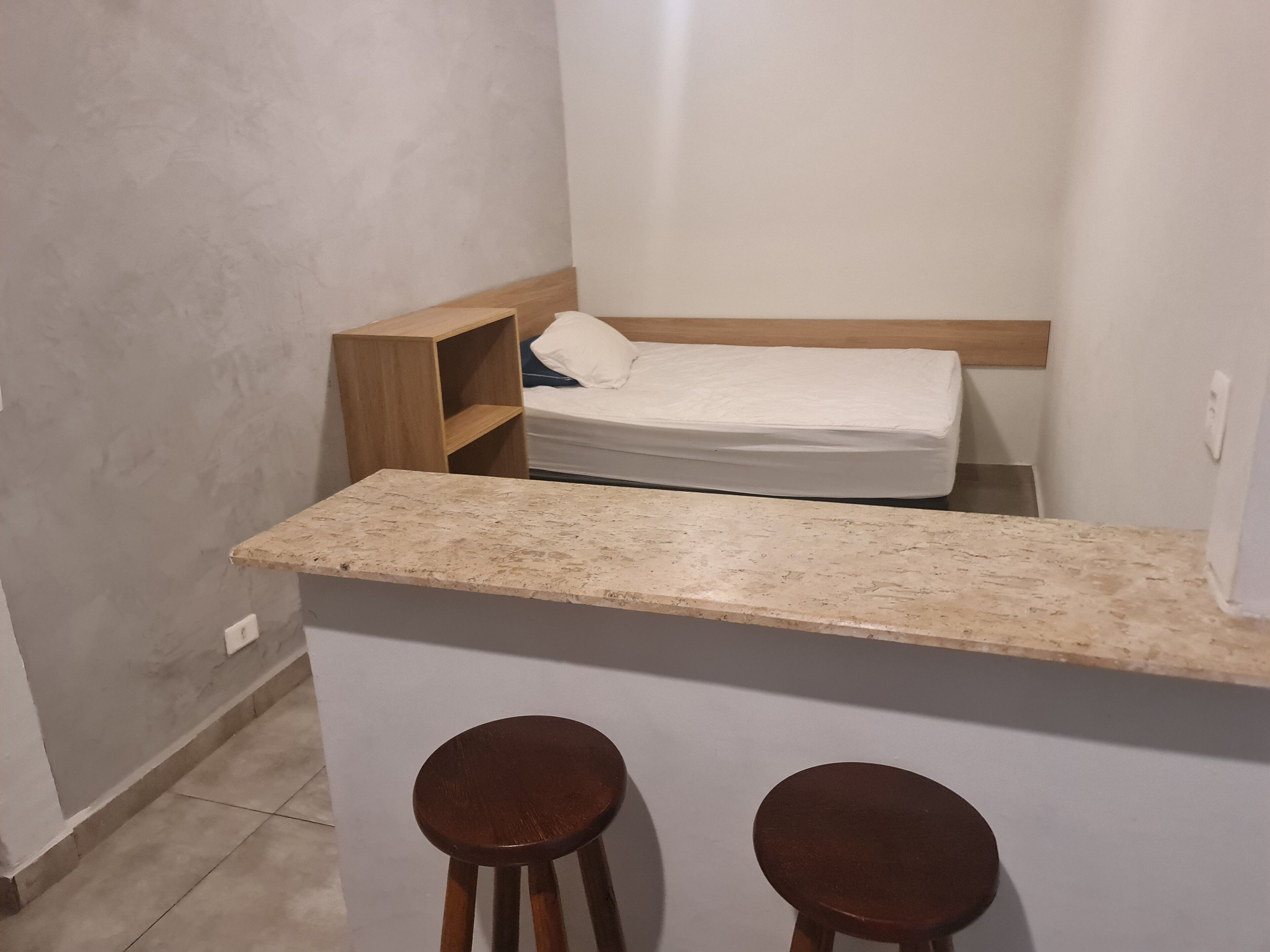 A small, compact room with a bed, a wooden shelf, and two stools.