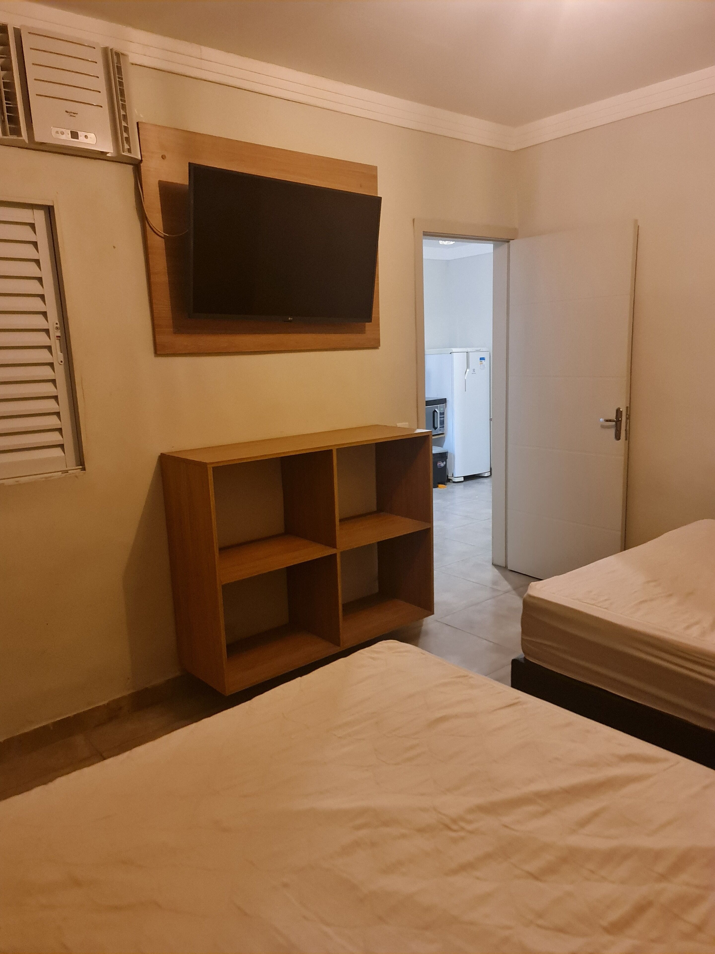 Quarto com duas camas, uma TV fixada na parede e uma estante de madeira.