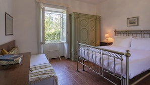 Ein Schlafzimmer mit Bett, Nachttisch, Lampe und Blick auf Grün hinter einem Fenster.