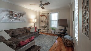 Un salon avec un canapé en cuir marron, un tapis bleu, un ventilateur de plafond, un téléviseur et une bibliothèque.