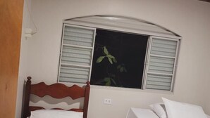 Ein kleines, sauberes Zimmer mit zwei Einzelbetten, einem Holz-Kopfteil, einem Fenster mit Jalousien und einem durch das Fenster sichtbaren Zimmerpflänzchen.
