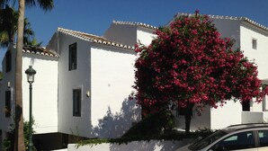 A white building with a balcony, a car parked in front, and a tree with pink flowers.