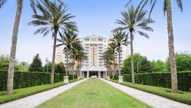 A hotel with a central entrance, surrounded by a symmetrical garden with palm trees and hedges.