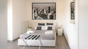A modern bedroom with a large bed, two bedside tables, a radiator, and a cityscape painting above the bed. - LUXURY 1 (Bari)