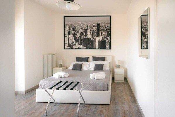 A modern bedroom with a large bed, two bedside tables, a radiator, and a cityscape painting above the bed. - LUXURY 1 (Bari)