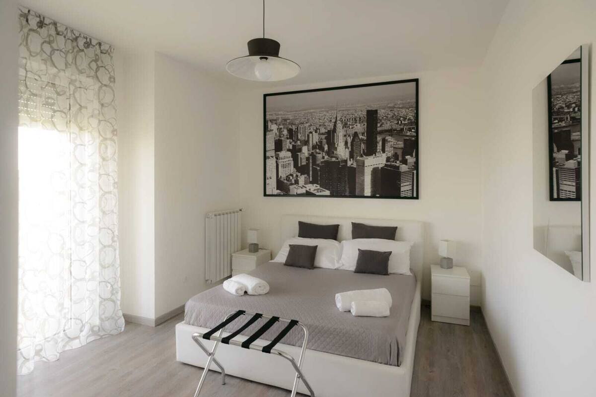 A modern hotel room with a large bed, two bedside tables, a radiator, and a cityscape artwork on the wall.