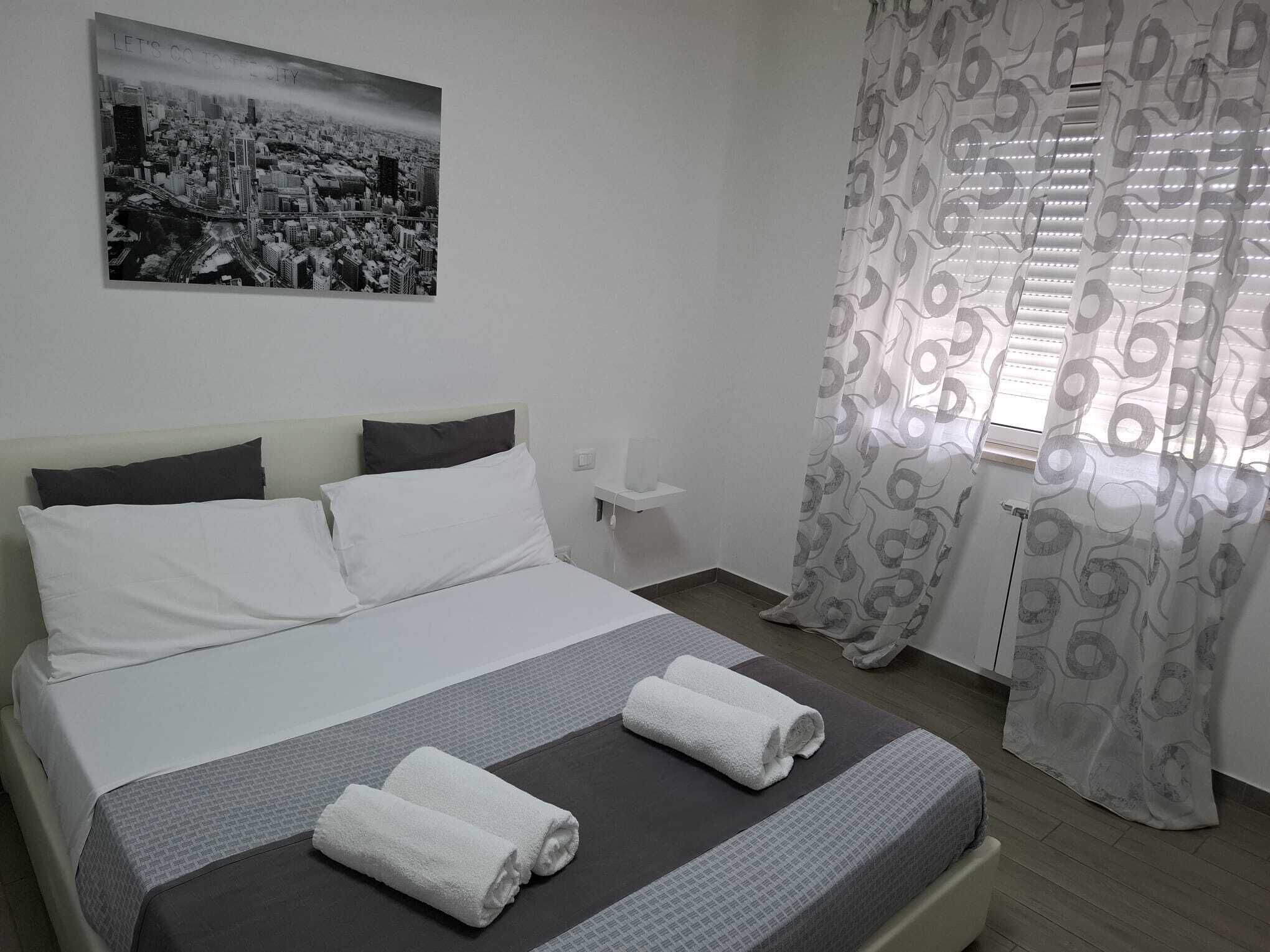 A neatly made bed with white and gray linens, a cityscape poster on the wall, and a patterned curtain.