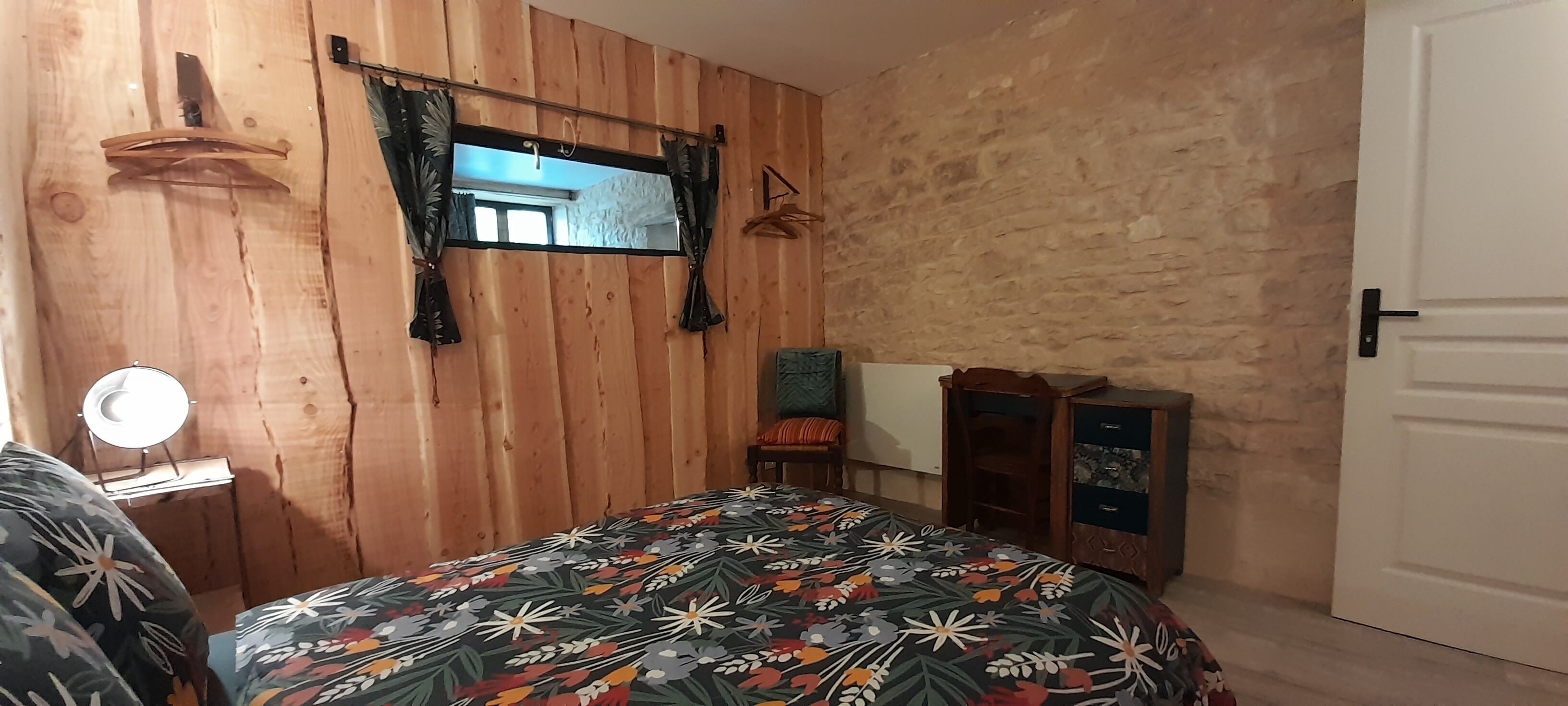 A bedroom with a wooden wall, a bed with a floral bedspread, a mirror, a chair, a small table with a lamp, and a wardrobe.