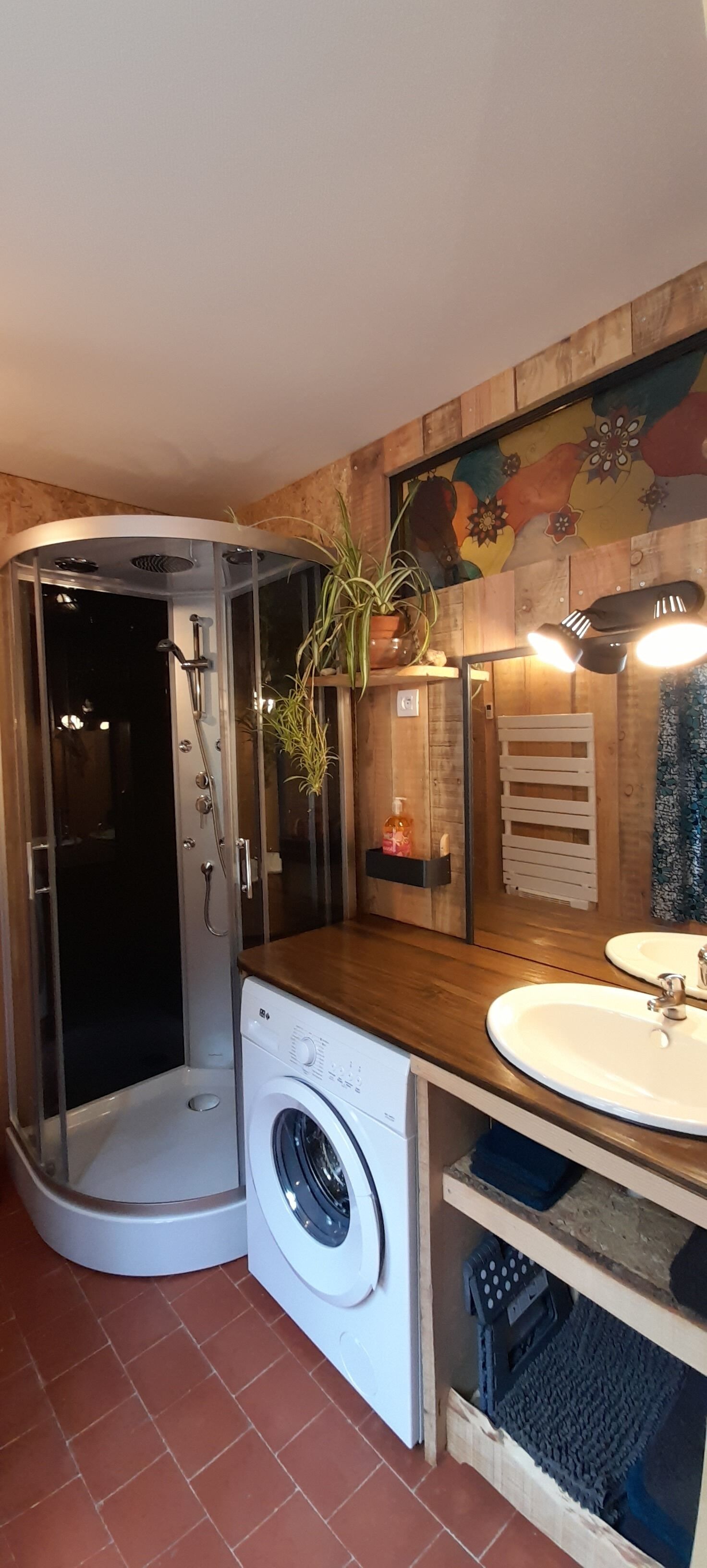 Une salle de bain compacte dotée d’une douche à parois de verre, d’un lave-linge encastré, d’un meuble-lavabo en bois et d’une fresque colorée sur le mur.