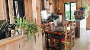 Interior de madera con mesa, sillas, zona de cocina y ventana con vistas a la vegetación.