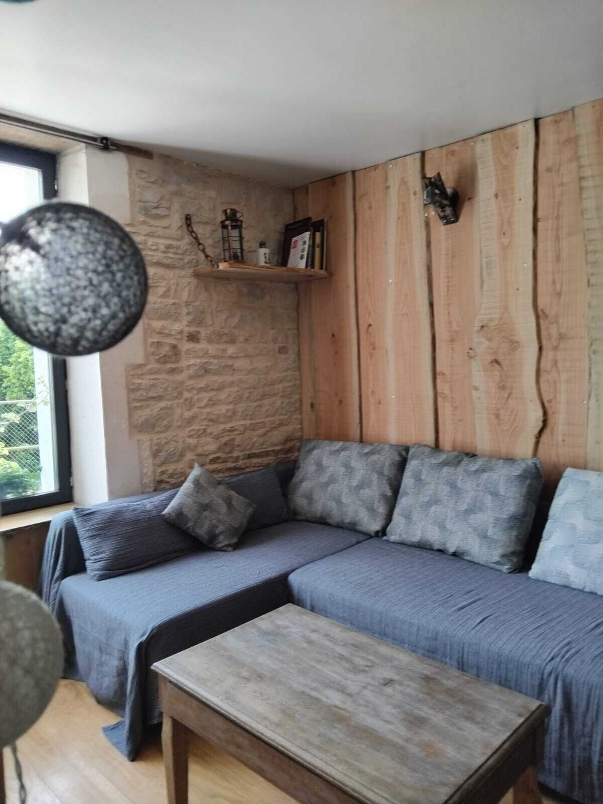 Una habitación acogedora con pared de piedra, paneles de madera, un sofá gris con cojines, una pequeña mesa de madera y una lámpara colgante.