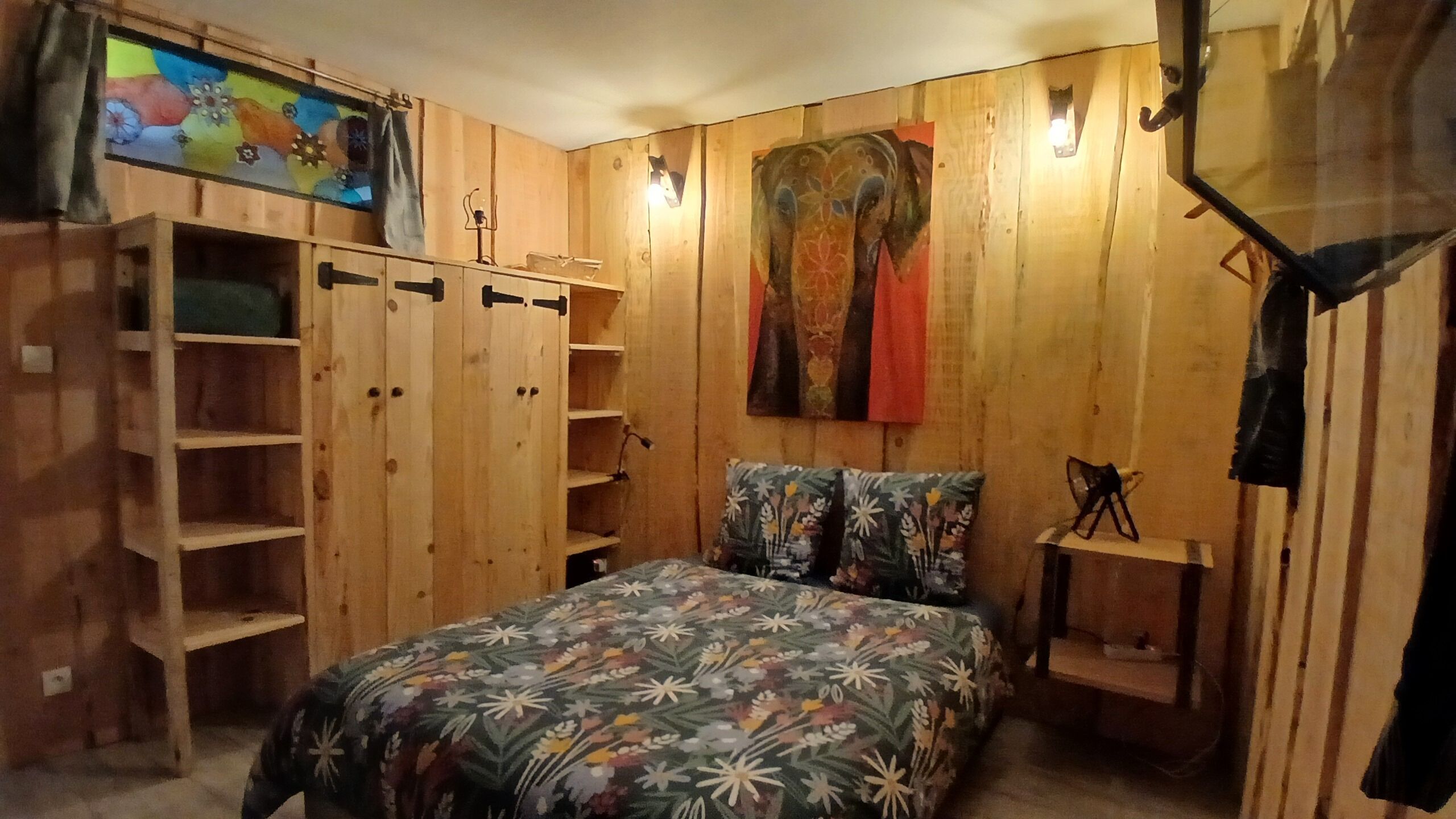 A bedroom with wooden walls, a bed with floral bedding, a shelf unit, and a painting on the wall.