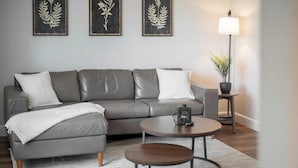 Un salon moderne avec un canapé gris, deux tables rondes et des œuvres d’art botanique encadrées accrochées au mur.