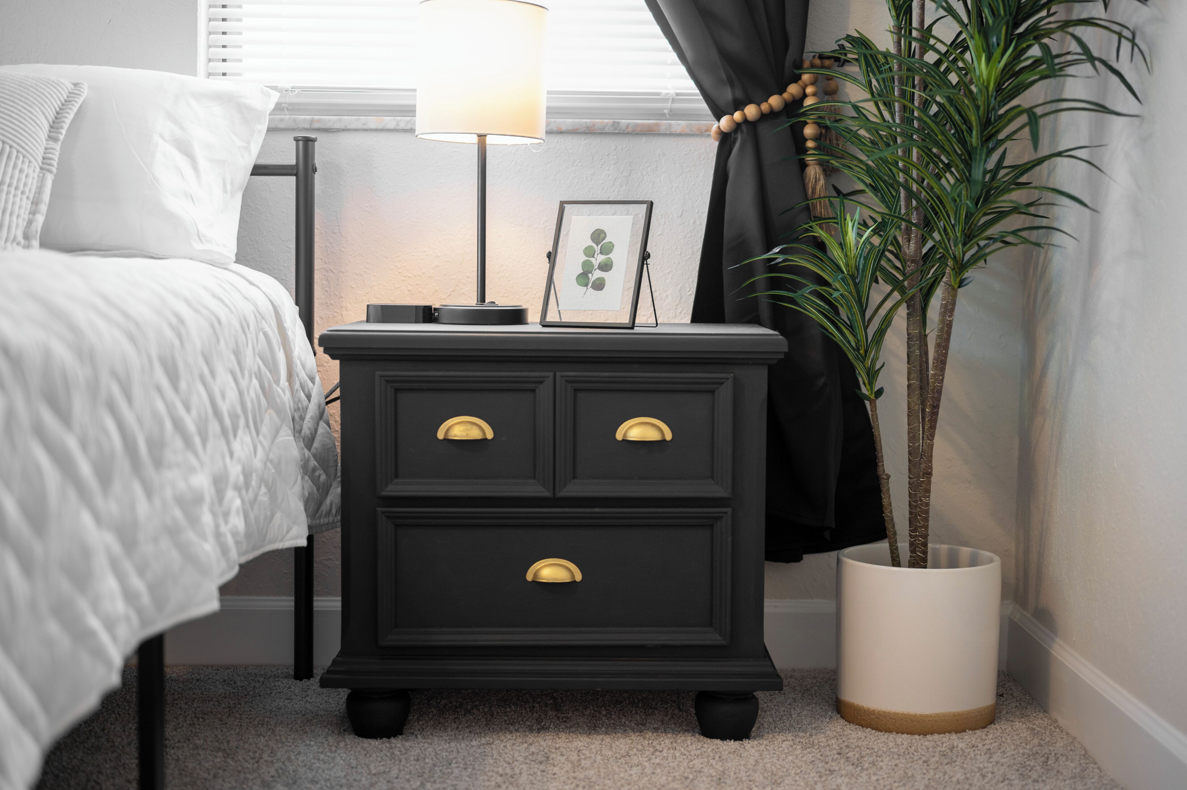 Une chambre avec une table de chevet noire, une lampe blanche, un tableau encadré et une plante en pot.