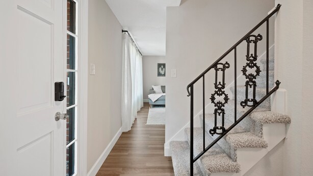 Un couloir avec une porte blanche, un escalier avec une rampe noire et un lit à l’arrière-plan.