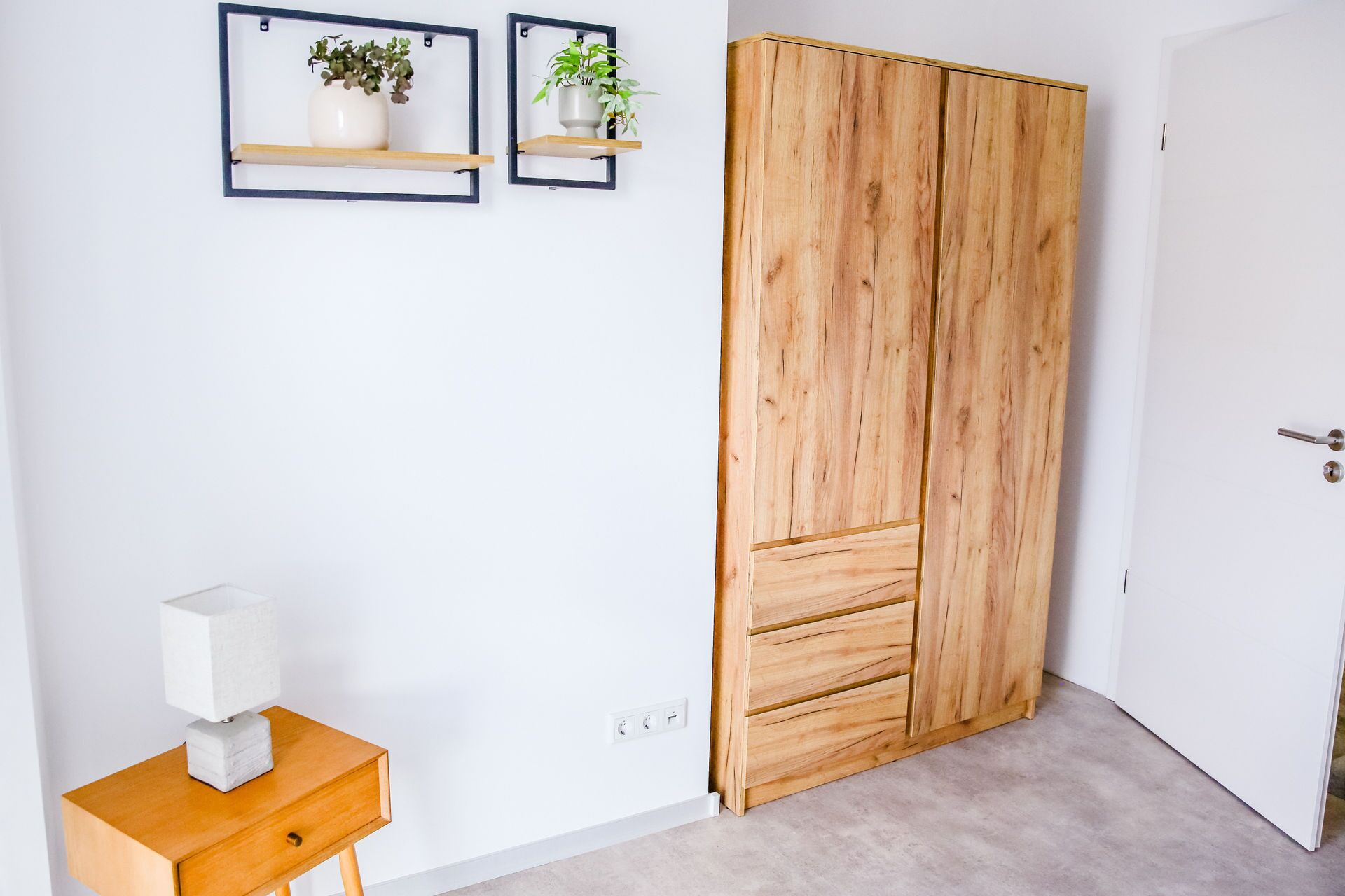 A room with a wooden wardrobe, a small wooden table with a lamp, and two wall-mounted shelves with plants.