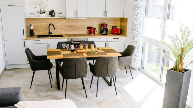 Una cocina moderna con armarios blancos, encimera de madera y un comedor con mesa y sillas. Un ventanal grande proporciona luz natural.